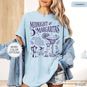 May include: Light blue t-shirt with the words "Midnight Margaritas" and a graphic of a margarita glass, moon, and other cocktail-related illustrations in purple. The shirt also has the text "More Color Options".