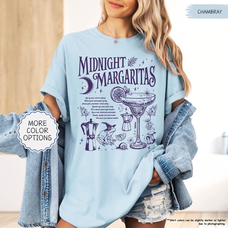 May include: Light blue t-shirt with the words "Midnight Margaritas" and a graphic of a margarita glass, moon, and other cocktail-related illustrations in purple. The shirt also has the text "More Color Options".