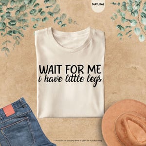 Pode incluir: Uma camiseta creme com o texto "WAIT FOR ME i have little legs" em preto. A camiseta est&aacute; estendida sobre uma superf&iacute;cie bege, com um par de jeans azuis e um chap&eacute;u marrom vis&iacute;veis.
