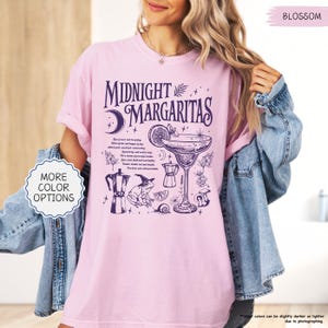 May include: A light pink t-shirt with the words "MIDNIGHT MARGARITAS" in dark purple, along with a cocktail illustration and other whimsical graphics. The shirt also has the text "MORE COLOR OPTIONS" in a white circle.