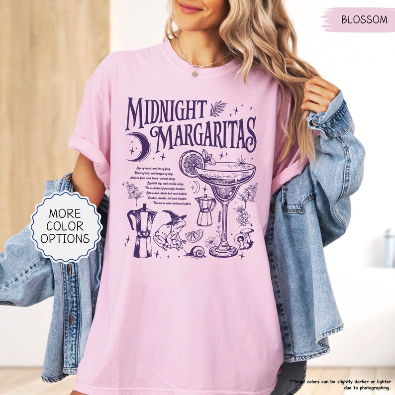 May include: A light pink t-shirt with the words "MIDNIGHT MARGARITAS" in dark purple, along with a cocktail illustration and other whimsical graphics. The shirt also has the text "MORE COLOR OPTIONS" in a white circle.