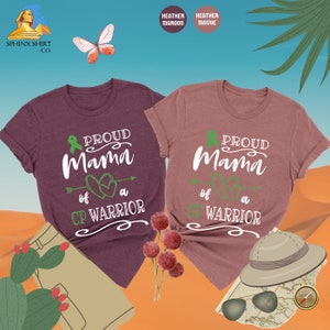 Op de afbeelding: Twee t-shirts in heidekleur met de tekst "Proud Mama of a CP Warrior" en een groen lint en een hartontwerp. De shirts liggen op een zandkleurige achtergrond.