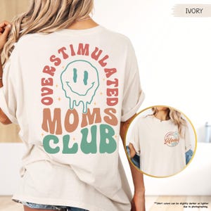 K&ouml;nnte beinhalten: Elfenbeinfarbenes T-Shirt mit dem Text "OVERSTIMULATED MOMS CLUB" in Retro-Schrift und einem schmelzenden Smiley-Gesicht. Das Shirt hat kurze &Auml;rmel und eine lockere Passform. Der Text "IVORY" befindet sich in der oberen rechten Ecke.
