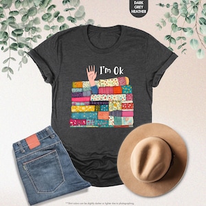 May include: Dark grey heather t-shirt with the text "I'm Ok" and a graphic of a hand and a stack of colorful patchwork quilts. The image also includes a pair of blue jeans and a tan hat.
