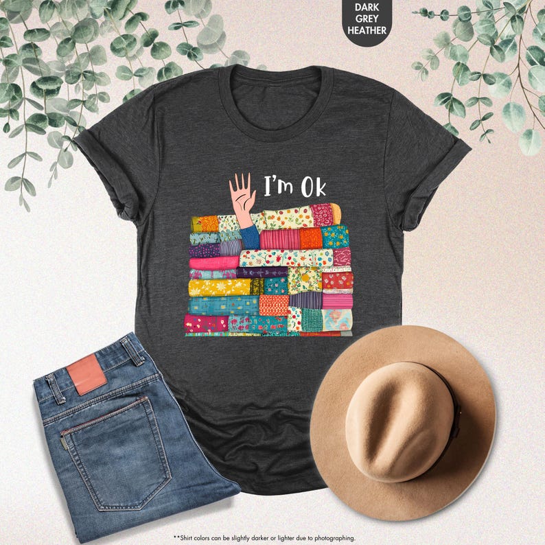 May include: Dark grey heather t-shirt with the text "I'm Ok" and a graphic of a hand and a stack of colorful patchwork quilts. The image also includes a pair of blue jeans and a tan hat.