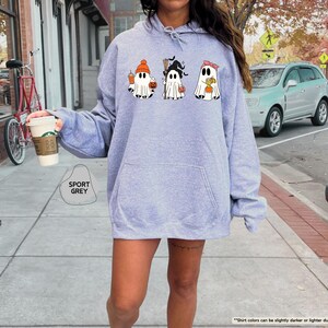 May include: A sport grey hooded sweatshirt featuring a graphic print of three white ghosts. The first ghost wears an orange beanie and holds a drink, the second wears a black witch hat and holds a broom, and the third wears a pink headband.