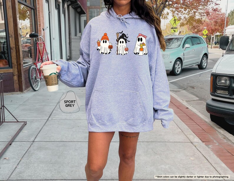 May include: A sport grey hooded sweatshirt featuring a graphic print of three white ghosts. The first ghost wears an orange beanie and holds a drink, the second wears a black witch hat and holds a broom, and the third wears a pink headband.