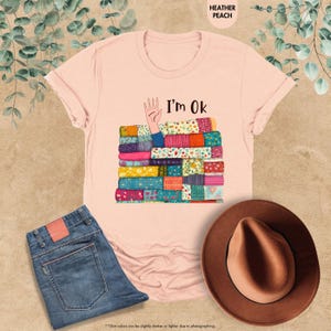 May include: Heather peach t-shirt with the text "I'm Ok" and a graphic of a hand and a stack of colorful, patterned fabric. The image also includes blue jeans and a brown hat.