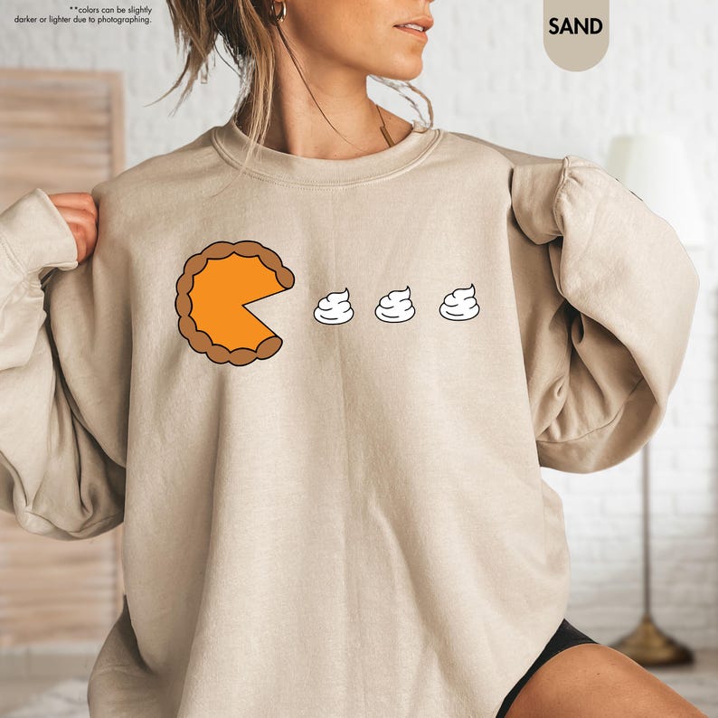May include: A beige sweatshirt with a graphic of a slice of pumpkin pie with a bite taken out of it, and three dollops of whipped cream. The text "SAND" is in the upper right corner of the image.