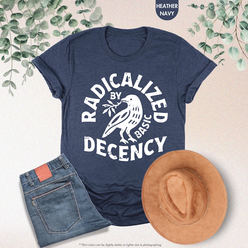 May include: A navy blue t-shirt with the words "RADICALIZED BY BASIC DECENCY" in a white, circular design. The design includes a dove holding a sprig. A pair of blue jeans and a brown hat are also in the image.