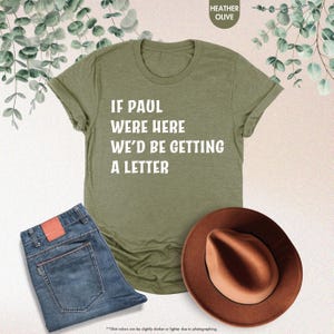 May include: An olive green t-shirt with the text "IF PAUL WERE HERE WE'D BE GETTING A LETTER" in white. The shirt is paired with blue jeans and a brown fedora hat. The image also includes eucalyptus branches.