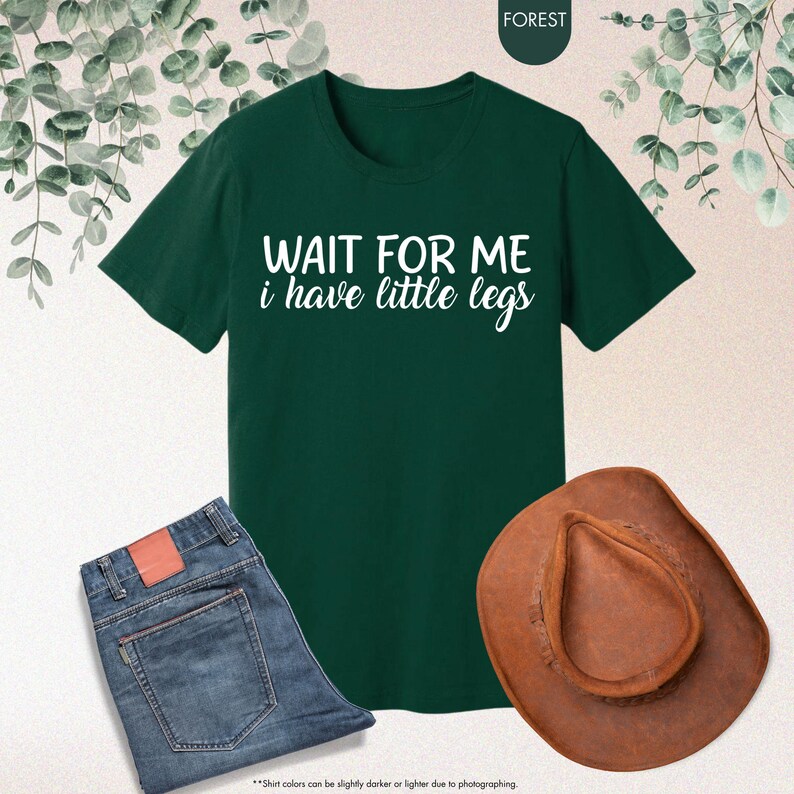 Pode incluir: Uma t-shirt verde escuro com o texto branco "WAIT FOR ME i have little legs". A imagem tamb&eacute;m inclui um par de jeans azuis e um chap&eacute;u de cowboy castanho. A t-shirt &eacute; uma pe&ccedil;a de roupa casual.