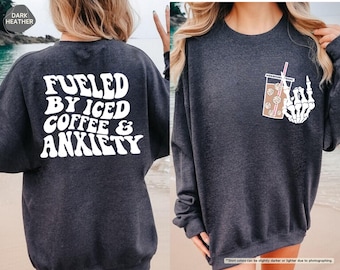May include: Dark heather grey sweatshirt with the text "Fueled by Iced Coffee & Anxiety" in white retro font on the back. The front of the sweatshirt features a white skeleton hand holding a cup of iced coffee with a straw.
