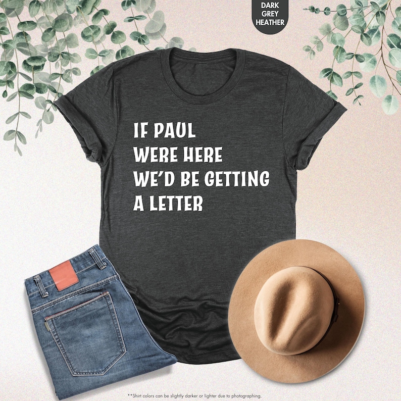 May include: Dark grey heather t-shirt with the text "IF PAUL WERE HERE WE'D BE GETTING A LETTER" in white. The shirt is paired with blue jeans and a tan hat. The image also includes a sprig of green leaves.