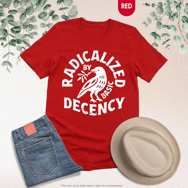 May include: A red t-shirt with the text "RADICALIZED BY BASIC DECENCY" in a circular design around a white bird holding a branch. The image also includes blue jeans and a beige hat.