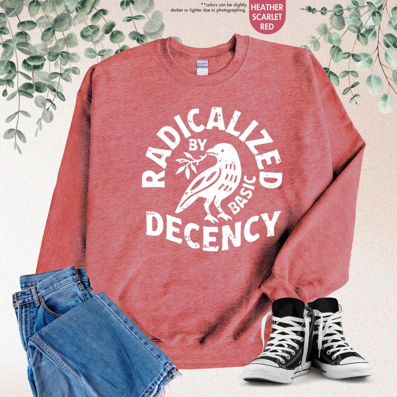 May include: Heather scarlet red sweatshirt with the text "RADICALIZED BY BASIC DECENCY" in a white circular design with a bird graphic. The sweatshirt is paired with blue jeans and black high-top sneakers.