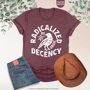 May include: A maroon t-shirt with the text "RADICALIZED BY BASIC DECENCY" and a white bird graphic. The image also includes a pair of blue jeans and a brown cowboy hat.