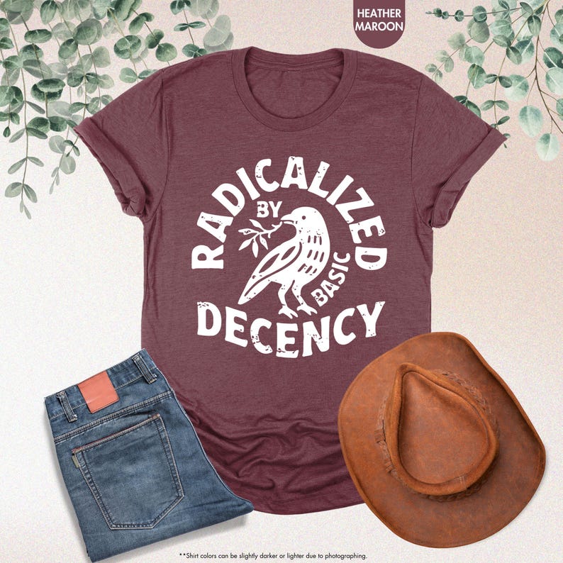 May include: A maroon t-shirt with the text "RADICALIZED BY BASIC DECENCY" and a white bird graphic. The image also includes a pair of blue jeans and a brown cowboy hat.