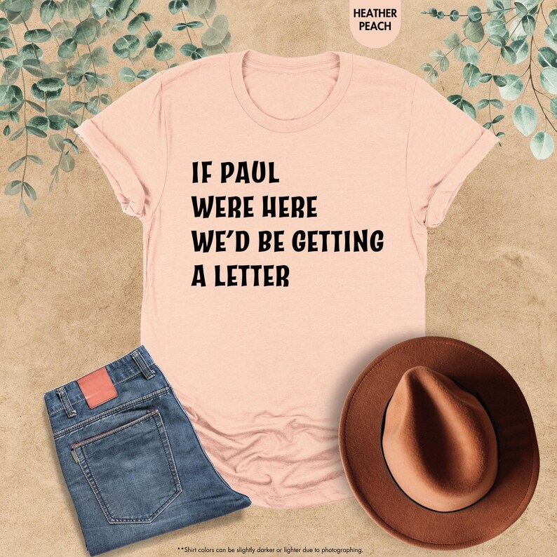 May include: A heather peach t-shirt with the text "IF PAUL WERE HERE WE'D BE GETTING A LETTER" in black. The shirt is paired with blue jeans and a brown hat. The image is taken from above.
