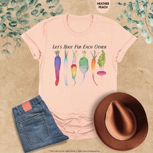 May include: A light pink t-shirt with the text "Let's Root For Each Other" and a watercolor illustration of colorful carrots, radishes, and turnips.