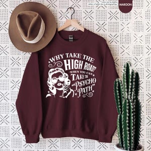 May include: Maroon sweatshirt with white graphic text and illustration. The text reads "WHY TAKE THE HIGH ROAD? WHEN YOU CAN TAKE THE 'PSYCHO PATH'". A brown hat hangs to the left. A cactus in a white pot is on the right.
