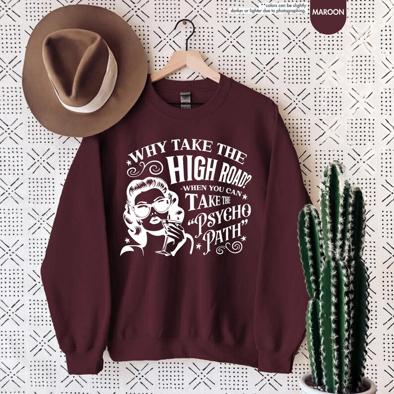 May include: Maroon sweatshirt with white graphic text and illustration. The text reads "WHY TAKE THE HIGH ROAD? WHEN YOU CAN TAKE THE 'PSYCHO PATH'". A brown hat hangs to the left. A cactus in a white pot is on the right.