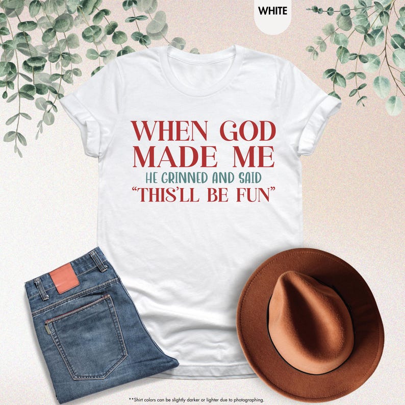 Puede incluir: Camiseta blanca con el texto rojo: "WHEN GOD MADE ME HE GRINNED AND SAID 'THIS'LL BE FUN'". La imagen tambi&eacute;n muestra unos vaqueros azules y un sombrero marr&oacute;n. La foto est&aacute; tomada desde arriba.