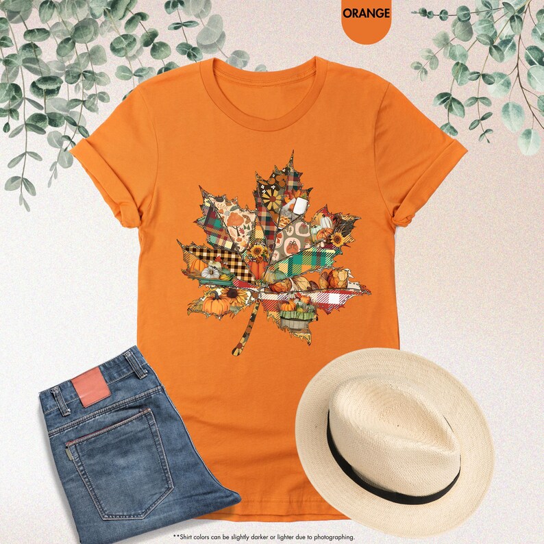 Puede incluir: Una camiseta naranja de manga corta con un gran gr&aacute;fico de hoja de arce compuesto por varios segmentos estampados que incluyen cuadros, calabazas, girasoles y follaje oto&ntilde;al. La prenda se muestra junto a unos vaqueros azules y un sombrero de paja.
