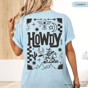 May include: A light blue t-shirt with a black graphic design featuring a cowboy riding a horse, a horseshoe, a cactus, and the word "HOWDY" in a western-style font.