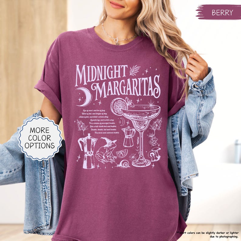 May include: Berry-colored t-shirt with the text "Midnight Margaritas" and a cocktail graphic. The design includes a crescent moon, a cocktail glass, and other illustrations. The shirt has rolled-up sleeves.
