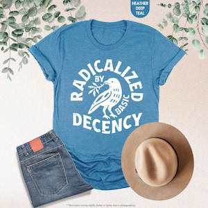 May include: A teal t-shirt with the text "RADICALIZED BY BASIC DECENCY" and a dove graphic. The image also shows a pair of blue jeans and a tan fedora hat. The shirt is folded at the sleeves.