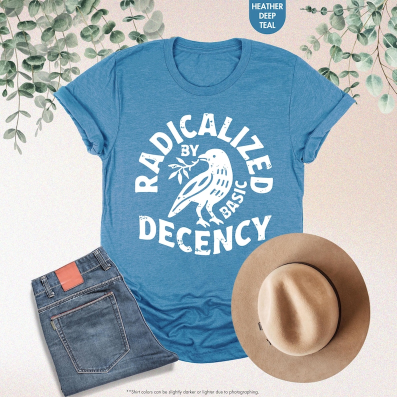 May include: A teal t-shirt with the text "RADICALIZED BY BASIC DECENCY" and a dove graphic. The image also shows a pair of blue jeans and a tan fedora hat. The shirt is folded at the sleeves.