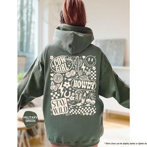 K&ouml;nnte beinhalten: Milit&auml;rgr&uuml;ner Kapuzenpullover mit einem grafischen Design mit den Worten "Cow Girl", "Howdy" und "Stay Wild". Das Design umfasst einen Kaktus, Stiefel, ein Hufeisen, eine Discokugel, ein Smiley und andere wei&szlig;e Elemente.