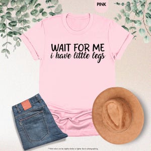 Pode incluir: Uma t-shirt rosa claro com o texto preto "WAIT FOR ME i have little legs". A imagem tamb&eacute;m mostra cal&ccedil;as de ganga azuis e um chap&eacute;u castanho. Folhas verdes adornam o fundo.