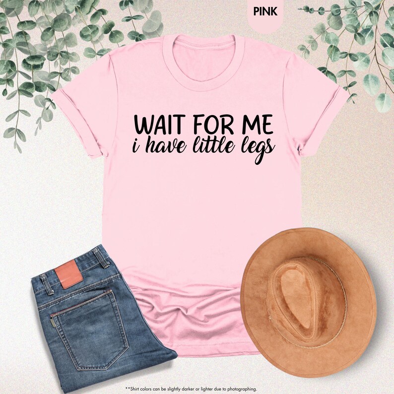 Pode incluir: Uma t-shirt rosa claro com o texto preto "WAIT FOR ME i have little legs". A imagem tamb&eacute;m mostra cal&ccedil;as de ganga azuis e um chap&eacute;u castanho. Folhas verdes adornam o fundo.