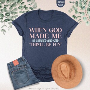 Puede incluir: Una camiseta azul marino con el texto "WHEN GOD MADE ME HE GRINNED AND SAID 'THIS'LL BE FUN'" en letras rosas. La camiseta se combina con unos vaqueros azules y un sombrero marr&oacute;n. Las palabras "Heather Navy" est&aacute;n en la parte superior.