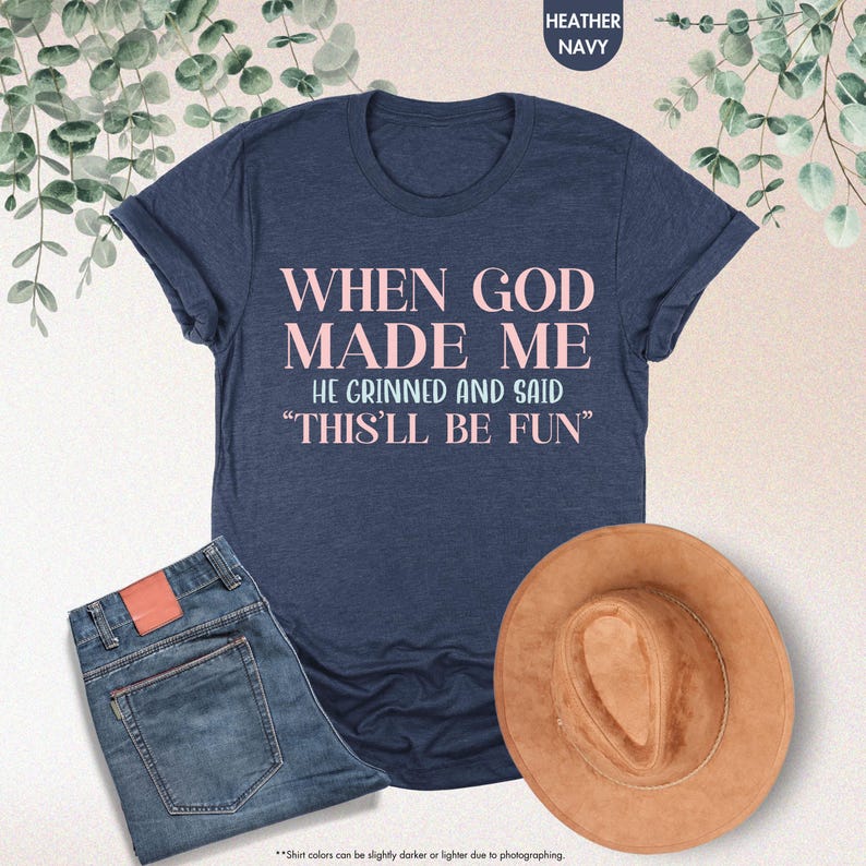 Puede incluir: Una camiseta azul marino con el texto "WHEN GOD MADE ME HE GRINNED AND SAID 'THIS'LL BE FUN'" en letras rosas. La camiseta se combina con unos vaqueros azules y un sombrero marr&oacute;n. Las palabras "Heather Navy" est&aacute;n en la parte superior.