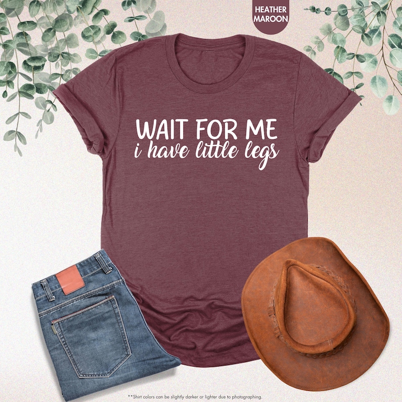 Pode incluir: T-shirt bord&ocirc; com o texto branco "WAIT FOR ME i have little legs". A imagem tamb&eacute;m inclui um par de jeans azuis e um chap&eacute;u de cowboy castanho.