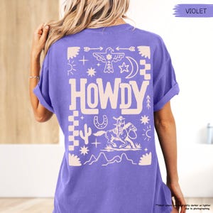 May include: A purple t-shirt with a white graphic design featuring the word "HOWDY" surrounded by western-themed symbols like stars, arrows, cacti, and a horseshoe.
