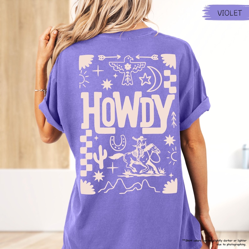 May include: A purple t-shirt with a white graphic design featuring the word "HOWDY" surrounded by western-themed symbols like stars, arrows, cacti, and a horseshoe.