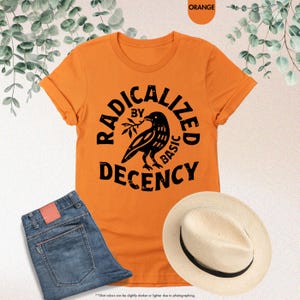 May include: Orange t-shirt with the text "RADICALIZED BY BASIC DECENCY" and a black bird graphic. The shirt is paired with blue jeans and a straw hat. The shirt is a crew neck style.