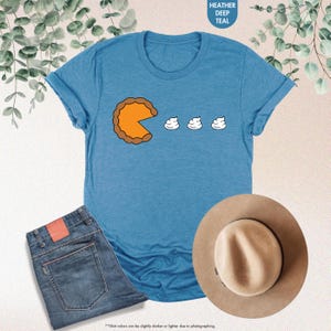 May include: A blue t-shirt with a graphic of a pumpkin pie slice eating three dollops of whipped cream. The text "HEATHER DEEP TEAL" is in the upper right corner of the image.
