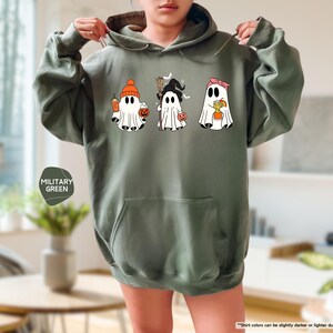 May include: A military green hooded sweatshirt featuring a graphic print of three white ghosts. The first ghost wears an orange beanie, the second a black witch hat with a broom, and the third a pink headband, each holding a small orange pumpkin bucket.