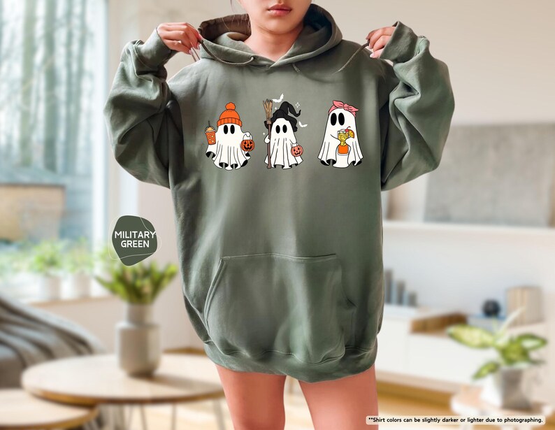 May include: A military green hooded sweatshirt featuring a graphic print of three white ghosts. The first ghost wears an orange beanie, the second a black witch hat with a broom, and the third a pink headband, each holding a small orange pumpkin bucket.