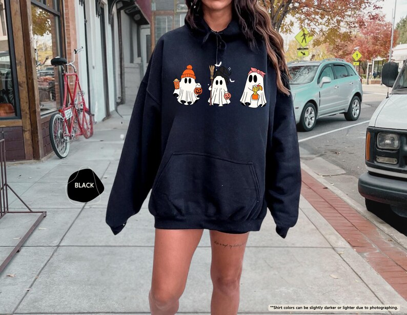 May include: A black hooded sweatshirt featuring a graphic print of three white ghosts. The first ghost wears an orange beanie, the second a black witch hat holding a broom, and the third a red headband. Each ghost holds a small orange pumpkin.