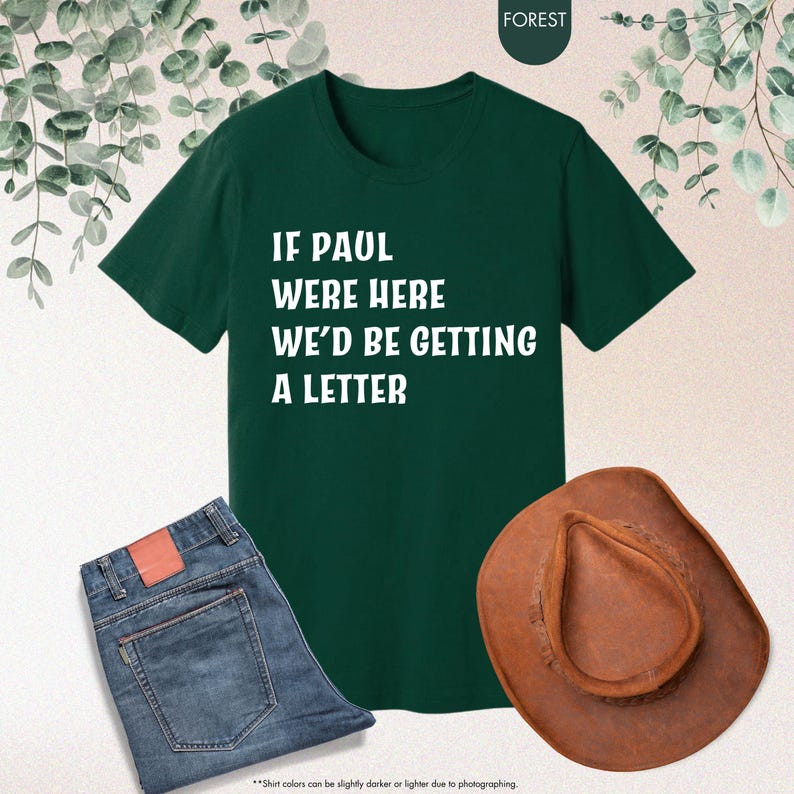 May include: A dark green t-shirt with the text "IF PAUL WERE HERE WE'D BE GETTING A LETTER" in white. The image also includes a pair of blue jeans and a brown cowboy hat. The shirt is the focal point.