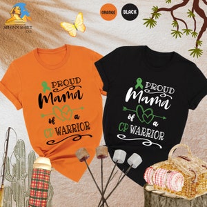 Op de afbeelding: Twee T-shirts, een oranje en een zwarte, met de tekst "Proud Mama of a CP Warrior" en een groen lint. De shirts worden getoond op een achtergrond met een picknickmand en een cactus.