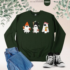 May include: A dark forest green crewneck sweatshirt featuring a graphic of three white ghosts. The first ghost wears an orange beanie and holds a coffee cup and pumpkin bucket. The second holds a broom and pumpkin. The third wears a pink headband.