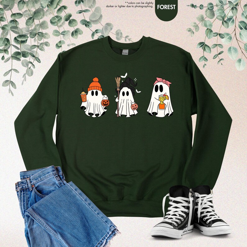 May include: A dark forest green crewneck sweatshirt featuring a graphic of three white ghosts. The first ghost wears an orange beanie and holds a coffee cup and pumpkin bucket. The second holds a broom and pumpkin. The third wears a pink headband.