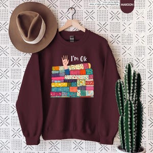 May include: Maroon sweatshirt with the text "I'm Ok" above a stack of colorful fabric. The fabric stack has a hand reaching out from it. A brown hat hangs above the sweatshirt.